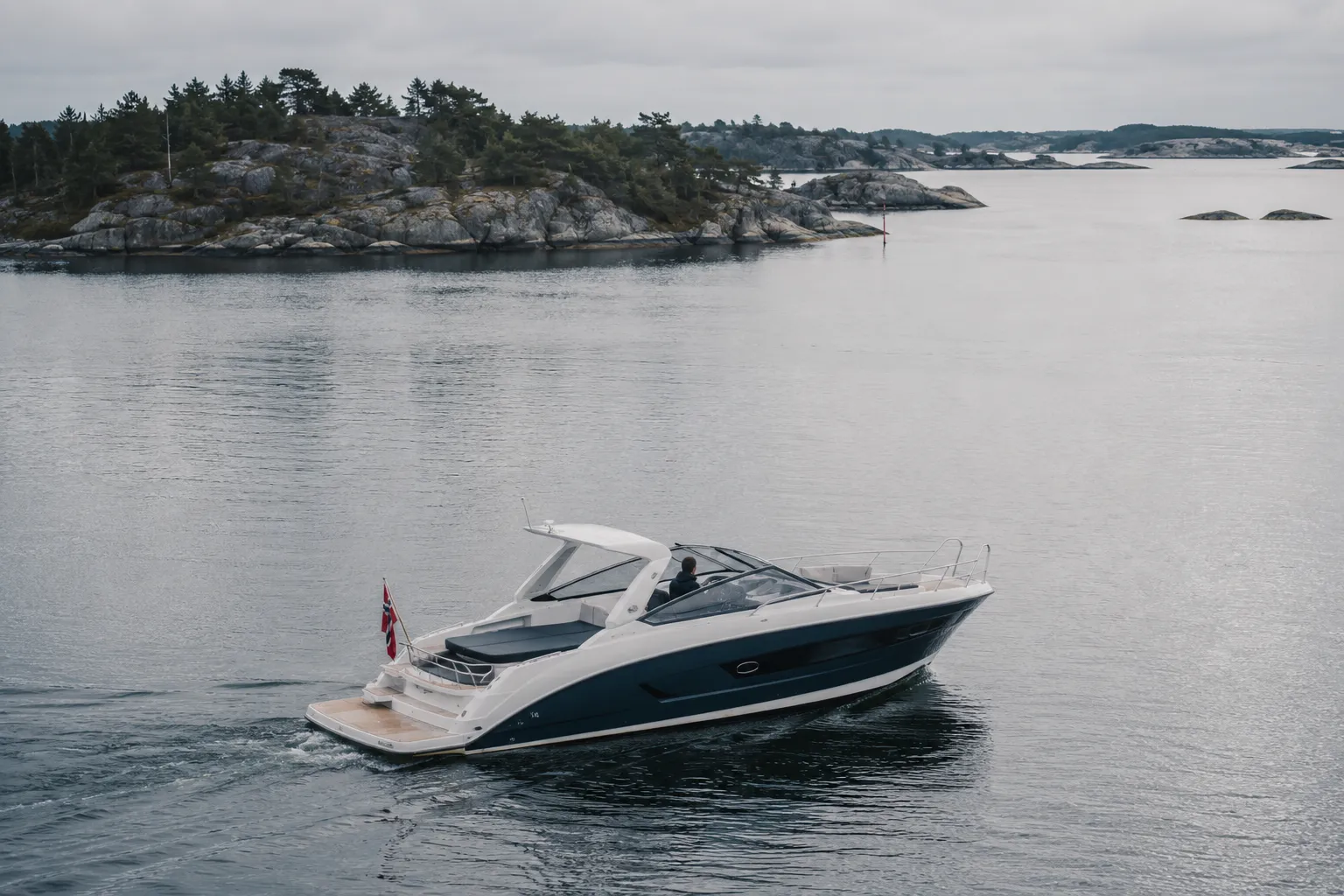 Moderne fritidsbåt på en rolig grå-blå fjord med skogkledd kyst i bakgrunnen.
