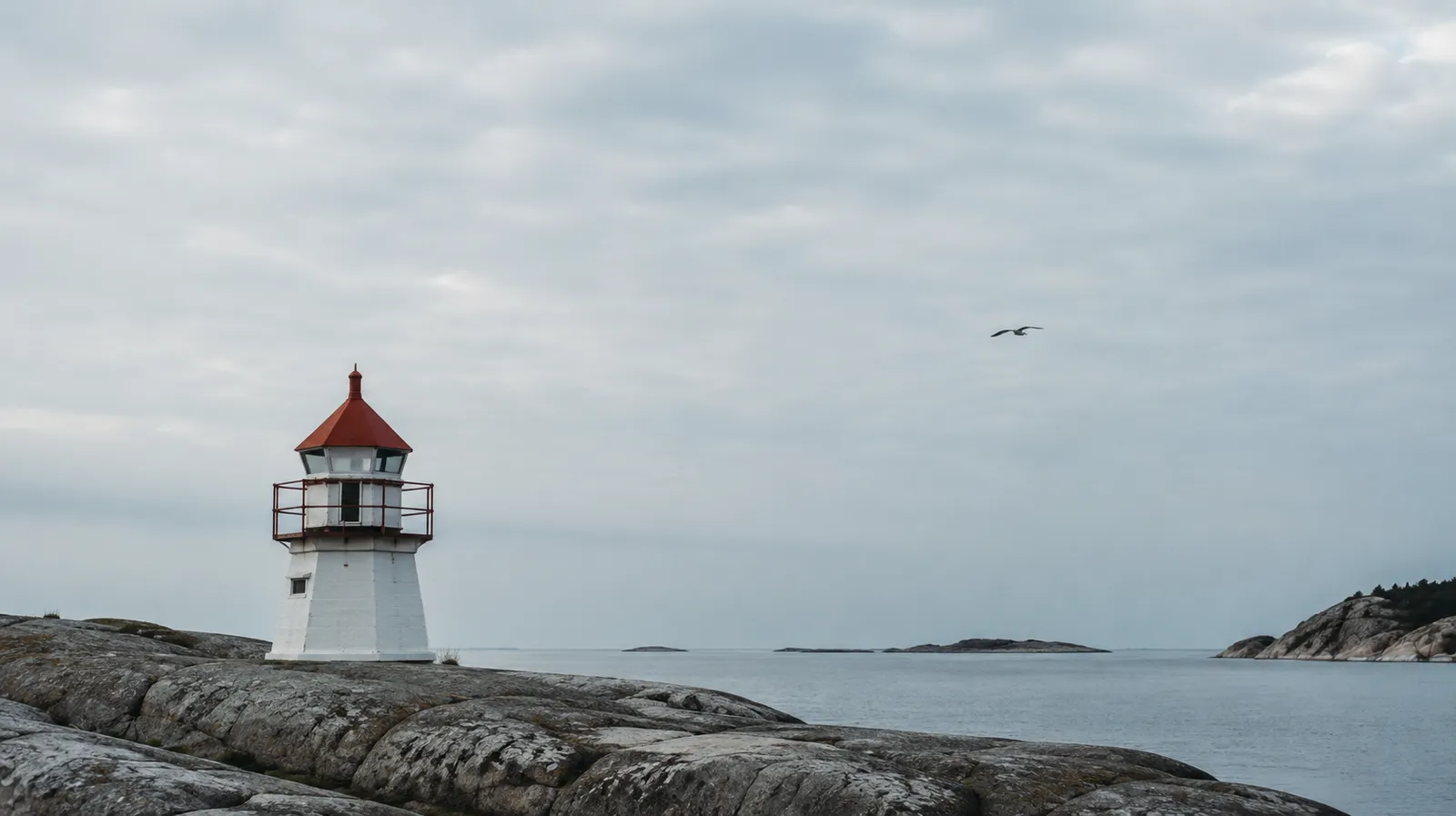 Liten norsk fyrlykt på nakne kystsvaberg med rolig sjø og overskyet himmel.
